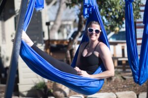 Anne, smiling, as she is seated in an aerial yoga hammock. 